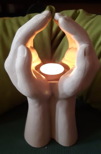 Ljusstake som visar två händer i böneställning. Ett tänt ljus finns i ljusstaken.